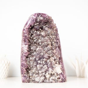 May include: A large amethyst geode crystal with a rough, textured surface. The crystal is a deep purple colour with white and grey inclusions. The geode is displayed on a white surface, with two white decorative objects in the background.