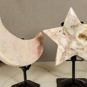 May include: Two decorative crystal sculptures, a crescent moon and a star, on black metal stands. The moon and star are both made of white and pink crystal with a natural, rough texture.