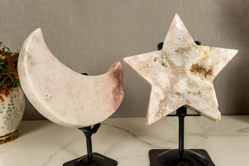 May include: Two decorative crystal sculptures, a crescent moon and a star, on black metal stands. The moon and star are both made of white and pink crystal with a natural, rough texture.