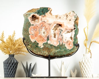 May include: A large, pink and green agate geode slice on a black metal stand. The geode has a natural, rough exterior and a smooth, polished interior with a variety of crystal formations.