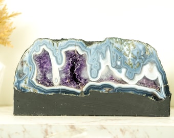 May include: A large amethyst geode with a natural, rough exterior and a polished interior. The geode is filled with purple amethyst crystals and has a white and blue agate banding pattern. The geode is displayed on a black base.