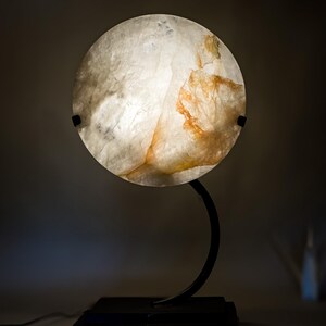 Pode incluir: Lâmpada redonda com abajur branco sujo e âmbar, suportada por uma estrutura de metal preto curvada. O abajur tem uma textura natural e a base é quadrada, criando um ambiente acolhedor.
