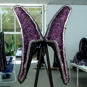 Op de afbeelding: Grote amethist geode sculpturen met een dieppaars kristallen interieur en een donker buitenframe. De geodes worden tentoongesteld op zwarte standaards, met andere amethiststukken zichtbaar op de achtergrond. Het algemene thema is natuurlijke kristallen en woondecoratie.