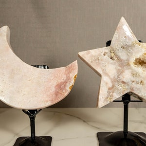 May include: Two pink crystal sculptures, a crescent moon and a five-pointed star, on black metal stands. The crystals have a white and pink colour with a natural, rough texture.