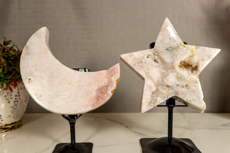 May include: Two pink crystal sculptures, a crescent moon and a five-pointed star, on black metal stands. The crystals have a white and pink colour with a natural, rough texture.