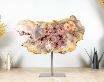 Könnte beinhalten: Eine große, unregelmäßig geformte Amethyst-Geode mit einer natürlichen, rauen Außenseite und einem hohlen Inneren, das mit Büscheln von rosa und violetten Kristallen gefüllt ist. Die Geode wird auf einem silbernen Metallständer präsentiert.