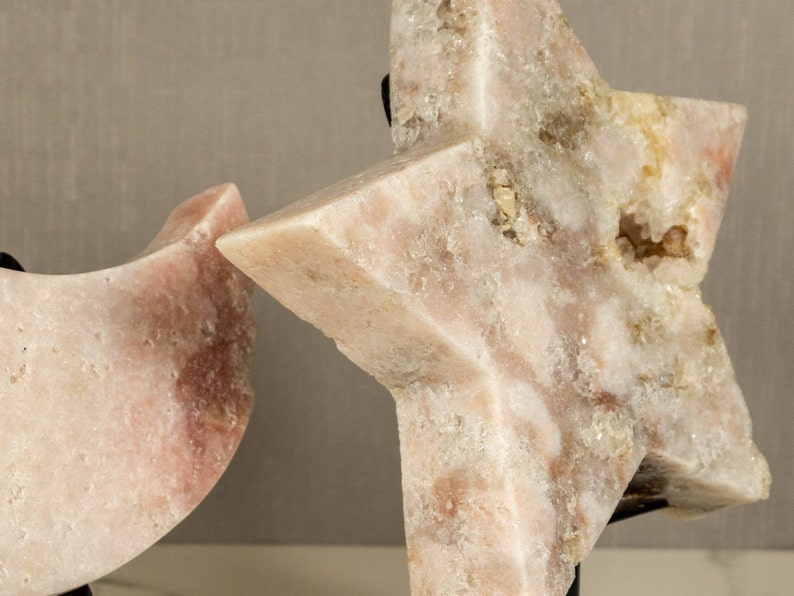 May include: Two pink crystal sculptures, one shaped like a crescent moon and the other a five-pointed star, are displayed on black stands.