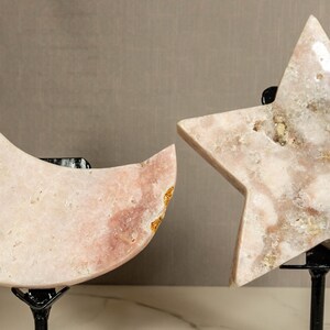 May include: Two pink crystal sculptures, a crescent moon and a five-pointed star, are displayed on black metal stands. The crystals have a natural, rough texture and are translucent.