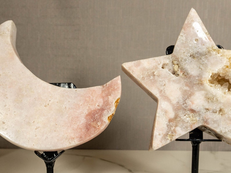 May include: Two pink crystal sculptures, a crescent moon and a five-pointed star, are displayed on black metal stands. The crystals have a natural, rough texture and are translucent.