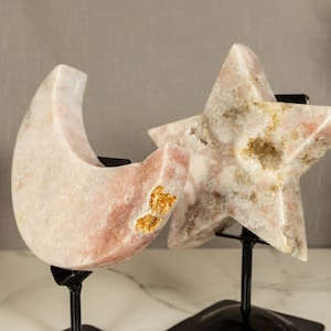 May include: Two pink crystal sculptures, a crescent moon and a five-pointed star, on black metal stands. The crystals have a white and gold marbled appearance.