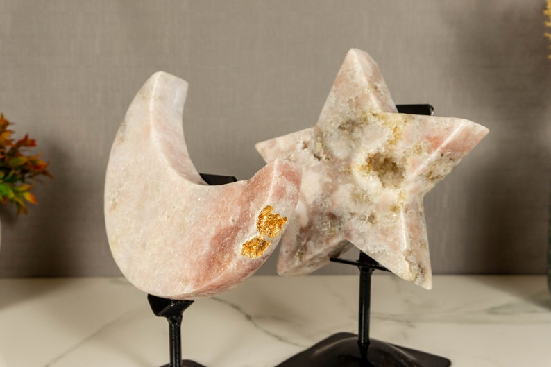 May include: Two pink crystal sculptures, a crescent moon and a five-pointed star, on black metal stands. The crystals have a white and gold marbled appearance.