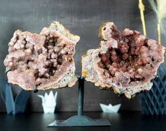 May include: Two pink and white geode halves with a crystal formation inside, displayed on a black metal stand.