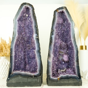 Op de afbeelding: Twee grote amethistgeoden met paarse kristallen erin. De geoden staan op een wit oppervlak met gedroogde bloemen in een vaas.