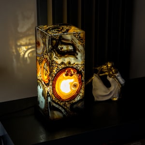 Pode incluir: Uma lâmpada retangular de ágata com um brilho amarelo quente. A lâmpada é feita de pedra de ágata natural com padrões marrons, brancos e laranja. A lâmpada está acesa e projeta uma sombra na superfície atrás dela.