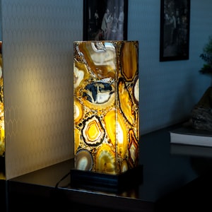 Pode incluir: Um abajur de mesa feito de ágata com um esquema de cores amarelo e marrom quente. A lâmpada está acesa e projeta um brilho quente. A lâmpada está sobre uma mesa marrom escura.