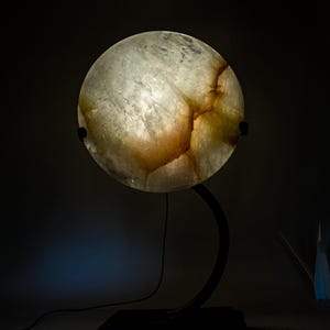 Pode incluir: Uma lâmpada redonda e iluminada com aparência de mármore. A lâmpada tem um suporte de metal escuro e curvo e uma base quadrada. A luz brilha através do material translúcido, revelando padrões marrons e brancos. O fundo é escuro.