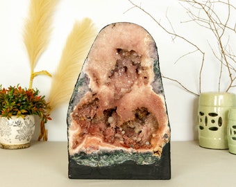 Könnte beinhalten: Eine große, rosafarbene Amethyst-Geode mit schwarzem Sockel. Die Geode ist mit Kristallen gefüllt und hat eine natürliche, raue Außenseite.