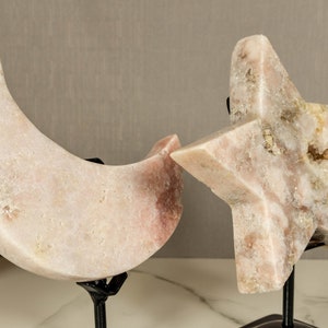 May include: A set of two pink crystal sculptures, one shaped like a crescent moon and the other like a star, both on black metal stands.
