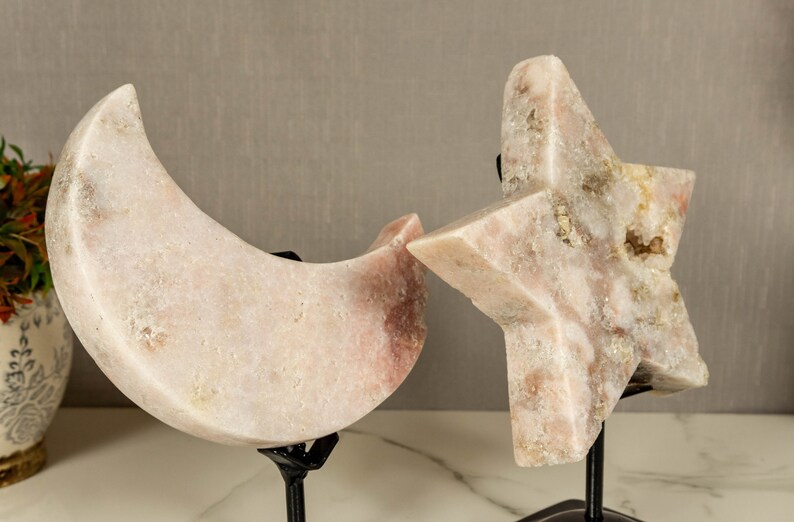 May include: A set of two pink crystal sculptures, one shaped like a crescent moon and the other like a star, both on black metal stands.