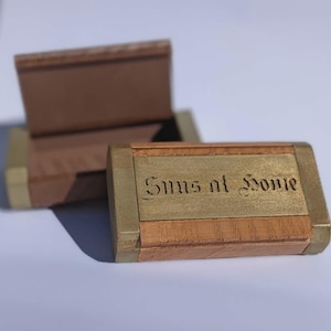 May include: Two wooden boxes with brass detailing. The front box features the words "Suns at Home" engraved in a decorative font. The boxes are rectangular with a hinged lid. The wood is light brown.
