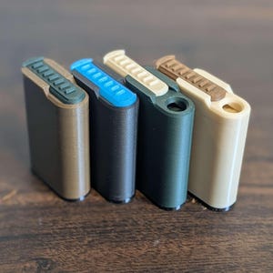 Puede incluir: Cuatro estuches rectangulares impresos en 3D en varios colores: marrón oscuro, negro, verde azulado y crema. Cada estuche tiene una parte superior de color contrastante con un diseño acanalado. Los estuches están dispuestos en fila sobre una superficie de madera.