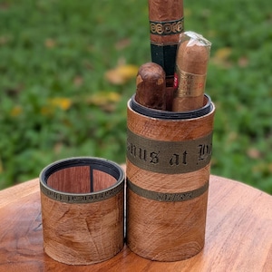 May include: A wooden cigar holder with the text "Venus at the Park" and the date "5/17/2020" engraved on it. The holder contains three cigars of varying sizes and colors. The holder is open, revealing the cigars inside.