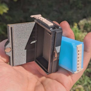 May include: A small, open, black box with a light brown top and a light blue rectangular object. The box has a textured surface and a hinged door. The light blue object has a light brown top and bottom.