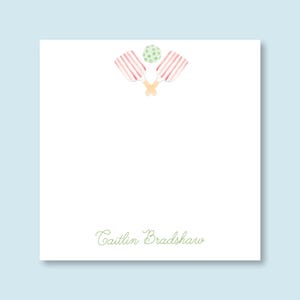 May include: A white card with a pink and white striped paddle ball set and a green ball in the upper left corner. The text "Caitlin Bradshaw" is in green cursive at the bottom of the card.