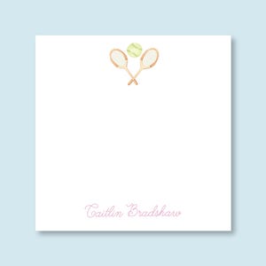 May include: White stationery card with a watercolor illustration of two crossed tennis rackets and a tennis ball. The name "Caitlin Bradshaw" is written in pink cursive at the bottom of the card.