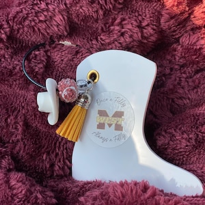 May include: A white, glossy, cowboy boot-shaped keychain with a gold-colored eyelet. The keychain features a small, yellow tassel, a pacifier charm, and a cluster of pink and silver beads. The text "Once a Filly, Always a Filly" is printed on the boot.