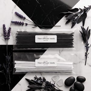 May include: Two packs of fiber diffuser reeds, one black and one white, are displayed on a marble surface. Each pack is labeled "en mer fiber diffuser reeds pack of 20." The image includes sprigs of lavender, dark leaves, and black stones.