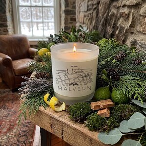 May include: A frosted glass candle with a lit wick, featuring a white label printed with a black line map and the text 'en mer MALVERN'. The candle is surrounded by evergreen branches, pinecones, green citrus fruits, moss, and lemon peels.