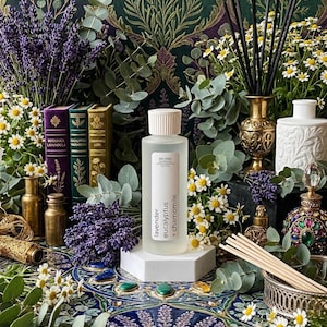 May include: A clear glass bottle with a white cap and a label that reads "en mer lavender eucalyptus + chamomile" sits on a white pedestal. The image is surrounded by lavender, eucalyptus, and chamomile flowers, with decorative bottles and books.