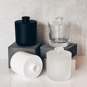 May include: Four cylindrical glass bottles in black, clear, white, and frosted white. The bottles have a rounded shape with a narrow neck and a small opening. The bottles are displayed on gray and white surfaces.