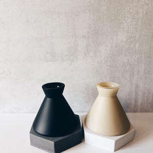 May include: Two ceramic candle holders, one black and one beige, with hexagonal bases. The candle holders are on a white surface.