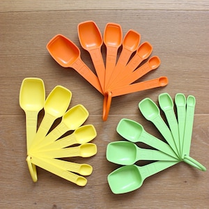 May include: Three sets of plastic measuring spoons in orange, yellow, and light green. Each set is held together by a ring at the base of the handles. The spoons vary in size, featuring clearly embossed measurement markings on the handles.