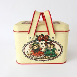 May include: A rectangular cream-colored metal tin with a red trim and two red hinged handles. The front features an oval illustration of two children in hats and dresses, framed by a lace-like border and a red bow with holly leaves and berries.