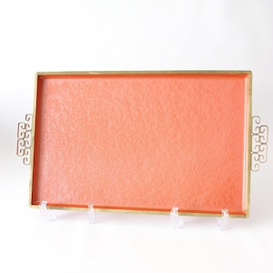 May include: Rectangular serving tray with a coral-colored interior and a gold-colored trim. The tray has decorative handles on each end and is displayed on clear stands. The tray is likely used for serving food or displaying items.