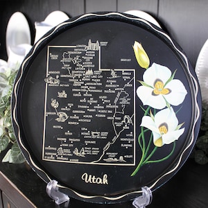 May include: A round black decorative tray featuring a gold-toned map of Utah with city names, landmarks, and the text Utah. The right side displays a painted illustration of white Sego Lily flowers with green stems and yellow centers.