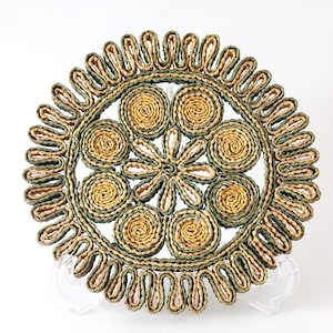 May include: A circular woven trivet featuring a central flower motif surrounded by eight coiled circles. The design uses alternating olive green and natural tan fibers, finished with a decorative scalloped edge made of looped green and tan cord.