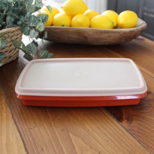May include: A rectangular food storage container with a white lid and a red base. The container is sitting on a wooden table. A wooden bowl filled with lemons is in the background.