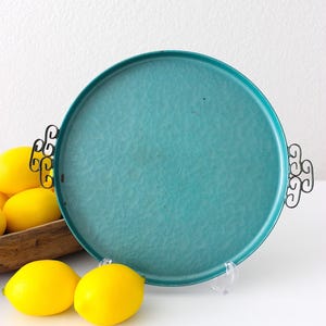 May include: A round, teal serving tray with decorative black handles. The tray has a textured surface and is displayed with several bright yellow lemons. The tray is likely used for serving food or displaying items.
