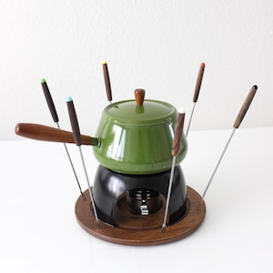 May include: Vintage fondue set with a green enamel pot, black base, and wooden serving tray. Includes several fondue forks with colored tips. The set is designed for tabletop cooking and serving.