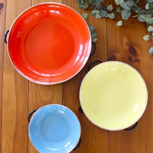 May include: Three colorful enamelware bowls in red, yellow, and blue, each with black handles and a silver rim. The bowls are arranged on a wooden surface, with a basket of greenery in the background.