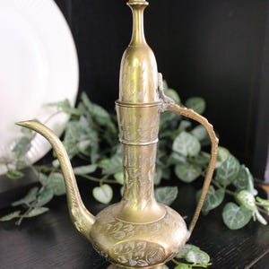 May include: A vintage brass water pitcher with an ornate design. The pitcher has a long, curved spout, a handle, and a decorative floral pattern. The pitcher is approximately 10 inches tall.