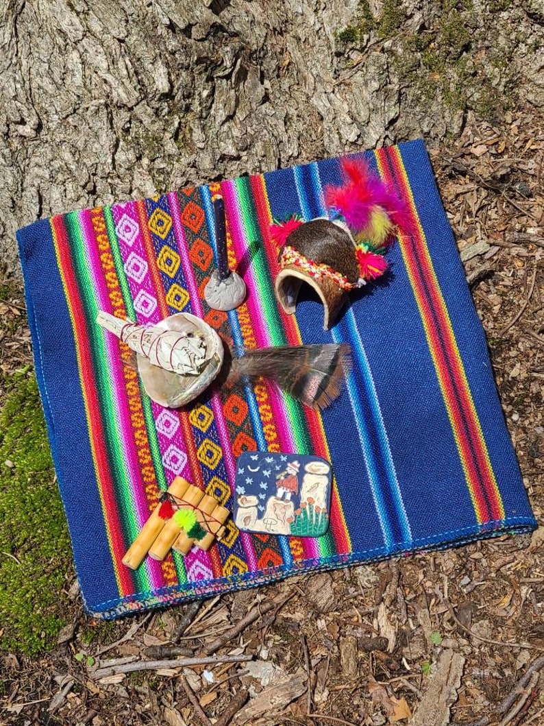 May include: A colorful woven blanket with a variety of spiritual items, including a seashell, a wooden pipe, a small drum, a feathered headdress, and a ceramic tile with a moon and stars design.
