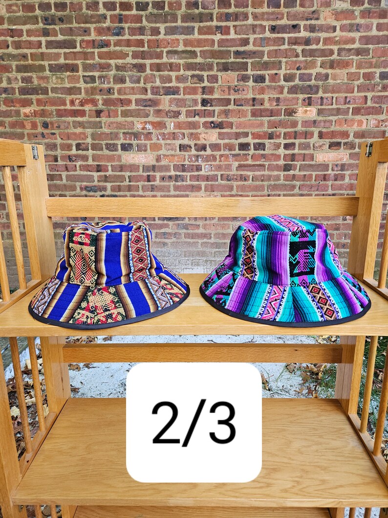 K&ouml;nnte beinhalten: Zwei bunte Bucket-Hats mit geometrischen Mustern. Der Hut links ist blau, lila und rot. Der Hut rechts ist lila, t&uuml;rkis und wei&szlig;. Die H&uuml;te befinden sich auf einem Holzregal mit der Nummer 2/3 auf einem wei&szlig;en Quadrat.