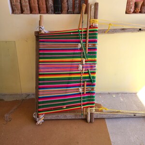 May include: A wooden loom with colorful yarn woven into a striped pattern. The loom is made of wood and has a simple design. The yarn is a variety of colors, including red, green, yellow, and blue.