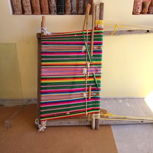 Puede incluir: Un telar de madera con hilo de colores tejido en un patr&oacute;n de rayas. El telar est&aacute; hecho de madera y tiene un dise&ntilde;o simple. El hilo es de varios colores, incluyendo rojo, verde, amarillo y azul.