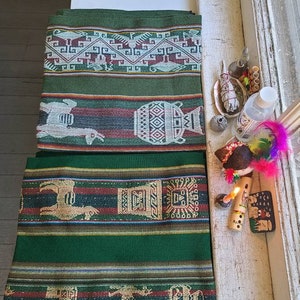 Puede incluir: Tres textiles tejidos en verde y marr&oacute;n con intrincados patrones, incluyendo llamas, dise&ntilde;os geom&eacute;tricos y un jarr&oacute;n estilizado. Los textiles est&aacute;n dispuestos sobre una superficie blanca con otros objetos, incluyendo una flauta de madera, una vela y una pluma.
